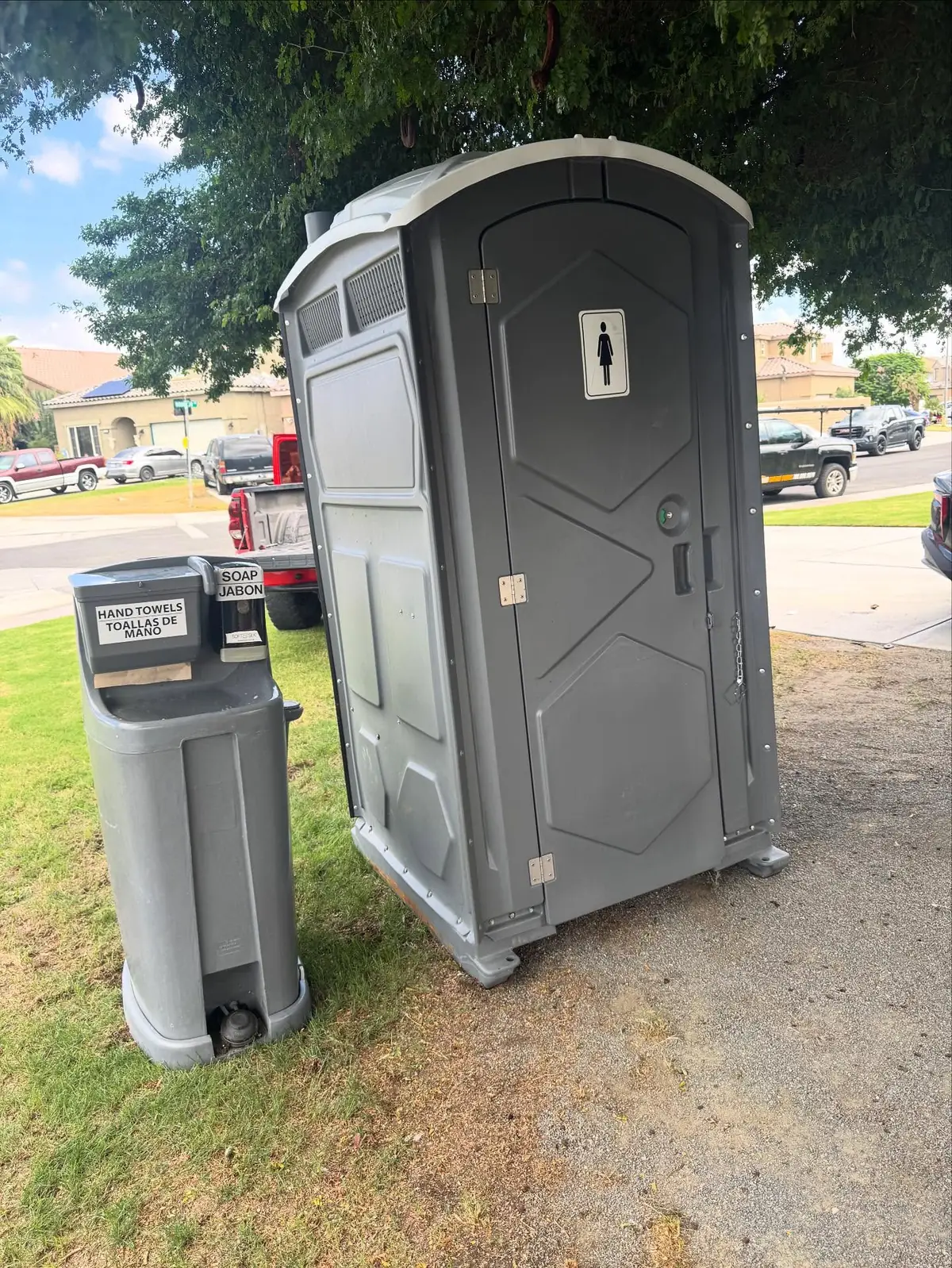 Foot-pump hand washing station standing next to a gray portable toilet on grass