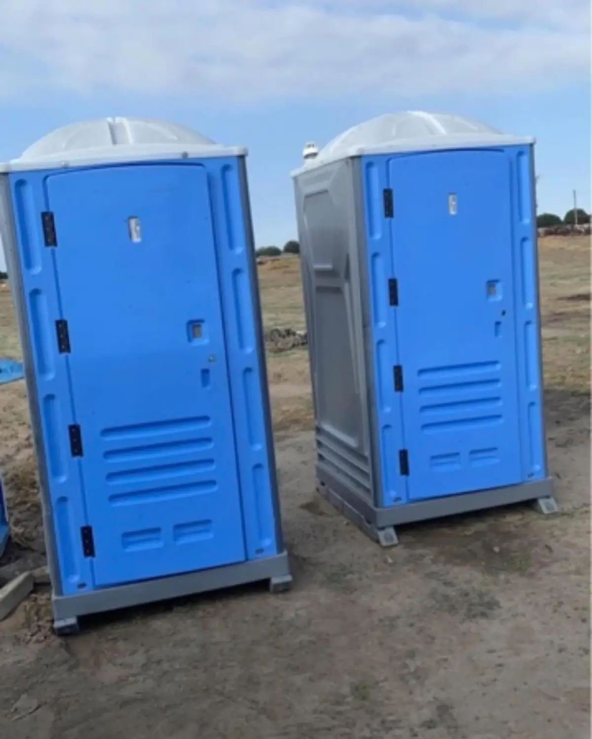 Standard portable toilets lined up on a gravel construction lot near a building under construction